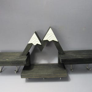 May include: A dark gray wooden wall shelf featuring a mountain-shaped design with white peaks. The unit includes three horizontal shelves at varying heights and seven silver metal hooks attached to the underside for hanging items.