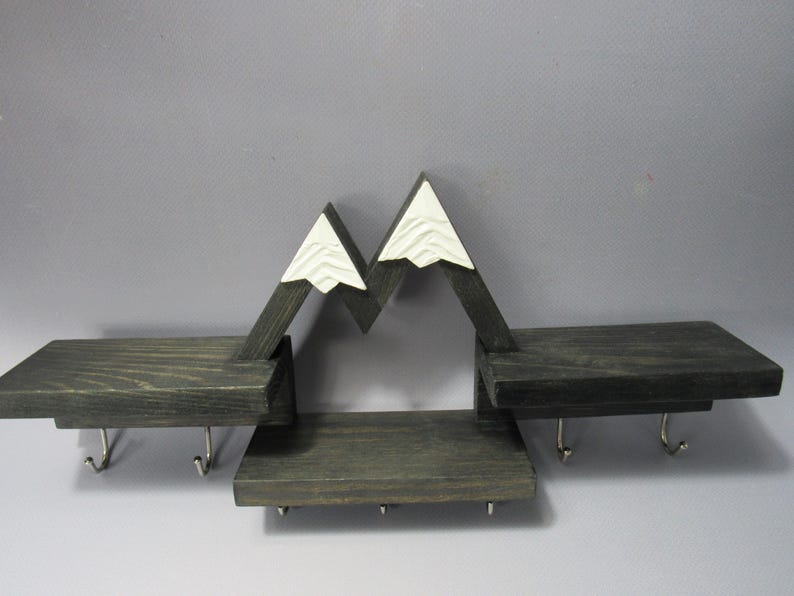May include: A dark gray wooden wall shelf featuring a mountain-shaped design with white peaks. The unit includes three horizontal shelves at varying heights and seven silver metal hooks attached to the underside for hanging items.