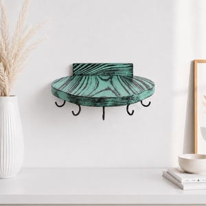 Puede incluir: Un estante de pared de madera en forma de semicírculo con un acabado desgastado en tonos verde azulado y negro. El estante incluye un panel trasero plano para el montaje y cinco ganchos de metal negro en la parte inferior para colgar objetos.