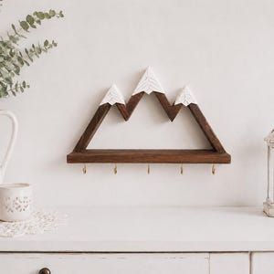 May include: A wall-mounted wooden shelf shaped like a mountain range with three peaks. The structure is made of dark brown wood with white painted tips on the peaks to resemble snow. Five small gold-colored metal hooks are attached to the bottom edge.