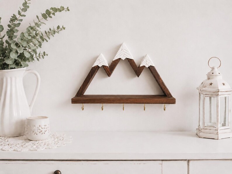 May include: A wall-mounted wooden shelf shaped like a mountain range with three peaks. The structure is made of dark brown wood with white painted tips on the peaks to resemble snow. Five small gold-colored metal hooks are attached to the bottom edge.