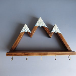 May include: Wooden mountain-shaped shelf with a brown stain and white painted peaks. The shelf has five hooks underneath for hanging items.