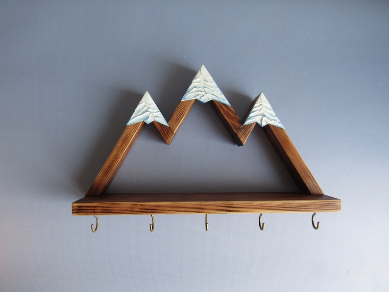 May include: Wooden mountain-shaped shelf with a brown stain and white painted peaks. The shelf has five hooks underneath for hanging items.