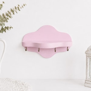 Puede incluir: Un estante de pared con un acabado en rosa suave y una silueta curva y festoneada. El estante cuenta con cuatro pequeños ganchos de metal oscuro unidos a la parte inferior, proporcionando un espacio funcional para colgar objetos.