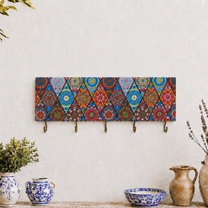 Puede incluir: Un perchero de pared rectangular con un vibrante patrón de diamantes multicolor que presenta intrincados motivos florales y geométricos. El tablero horizontal incluye cinco ganchos de metal color latón espaciados.