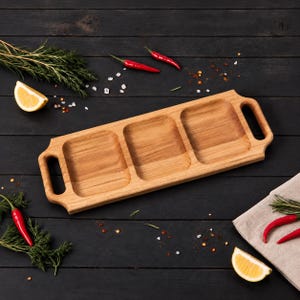 Puede incluir: Una bandeja de servicio rectangular de madera con tres compartimentos cuadrados distintos y dos asas integradas en los extremos. La bandeja está hecha de madera de tono claro con una veta visible, diseñada para organizar y servir alimentos.