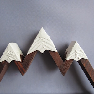 May include: A mountain-shaped wall decor piece crafted from dark brown stained wood. The three peaks are topped with textured, off-white material that mimics snow, featuring visible brushstrokes and a raised, layered surface finish.