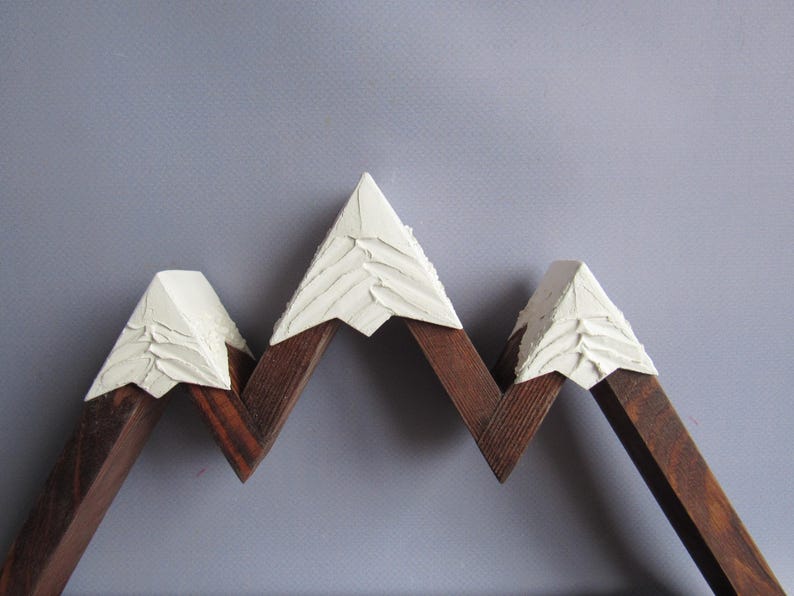 May include: A mountain-shaped wall decor piece crafted from dark brown stained wood. The three peaks are topped with textured, off-white material that mimics snow, featuring visible brushstrokes and a raised, layered surface finish.