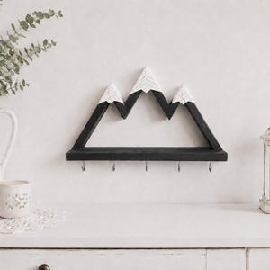 May include: A wall-mounted wooden shelf shaped like a mountain range with three peaks. The mountain structure is painted black with white, textured snow-capped tops. A flat black shelf base features five silver metal hooks for hanging items.