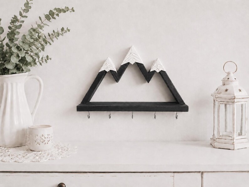 May include: A wall-mounted wooden shelf shaped like a mountain range with three peaks. The mountain structure is painted black with white, textured snow-capped tops. A flat black shelf base features five silver metal hooks for hanging items.