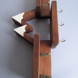 May include: A wall-mounted wooden key rack featuring a mountain-shaped design with dark brown stained wood and white painted peaks. The structure includes five small brass-colored metal hooks attached to the wooden frame for hanging items.