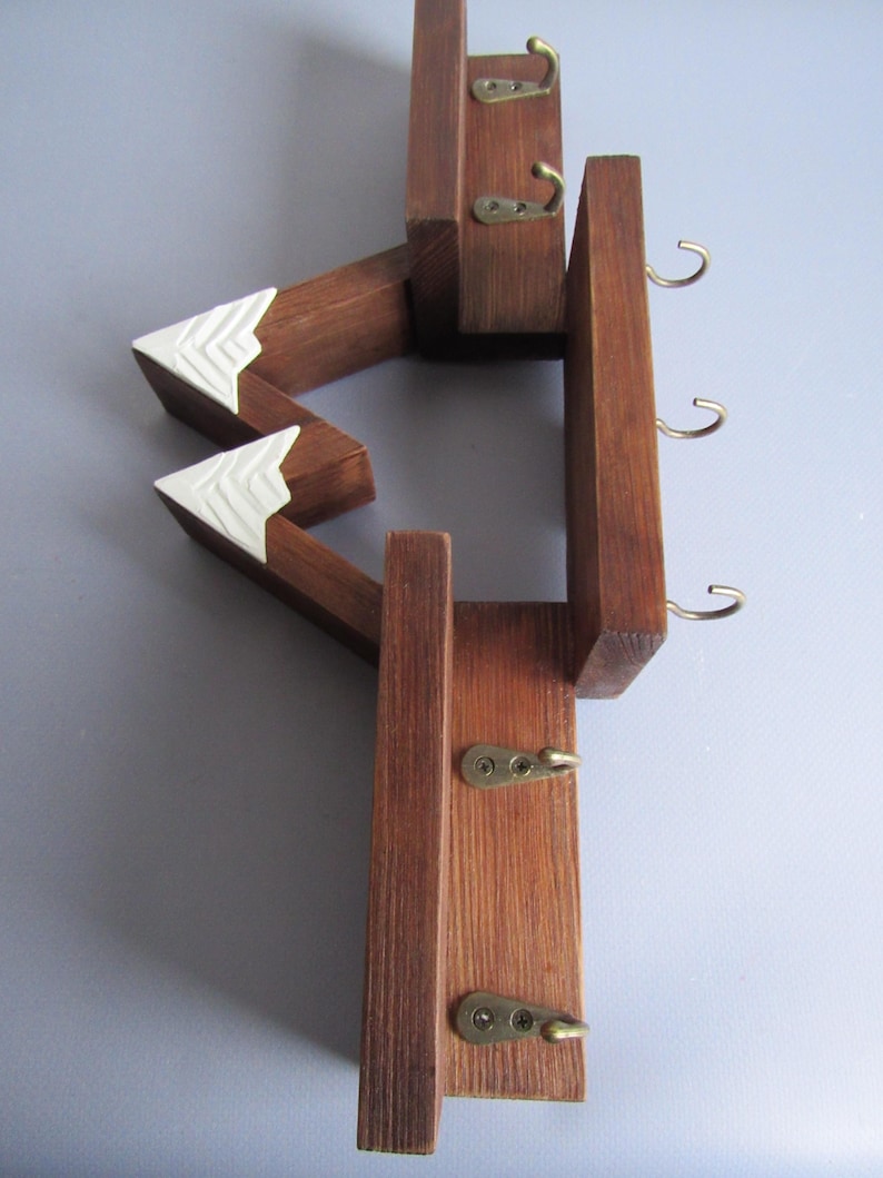 May include: A wall-mounted wooden key rack featuring a mountain-shaped design with dark brown stained wood and white painted peaks. The structure includes five small brass-colored metal hooks attached to the wooden frame for hanging items.
