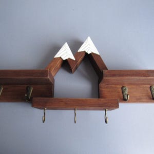 May include: A wall-mounted wooden shelf unit featuring a mountain-shaped design with two white-painted peaks. The dark brown wood structure includes three horizontal shelves and seven brass-colored metal hooks for hanging items.