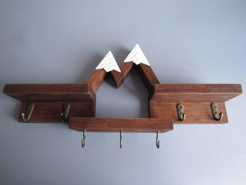 May include: A wall-mounted wooden shelf unit featuring a mountain-shaped design with two white-painted peaks. The dark brown wood structure includes three horizontal shelves and seven brass-colored metal hooks for hanging items.