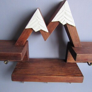 May include: A wall-mounted wooden shelf unit featuring a mountain range design with dark brown stained wood and white painted peaks. The structure includes three horizontal shelves and several small metal hooks attached to the underside.