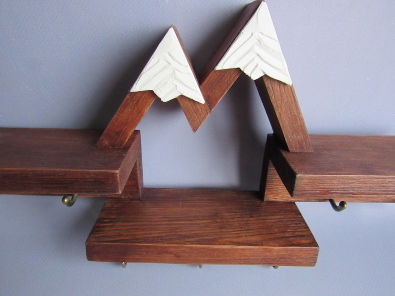 May include: A wall-mounted wooden shelf unit featuring a mountain range design with dark brown stained wood and white painted peaks. The structure includes three horizontal shelves and several small metal hooks attached to the underside.