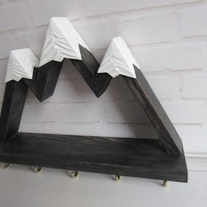 May include: A wall-mounted wooden shelf featuring a mountain range silhouette with dark charcoal stained wood bases and white painted peaks. The shelf includes five small gold-toned metal hooks attached to the bottom edge for hanging items.