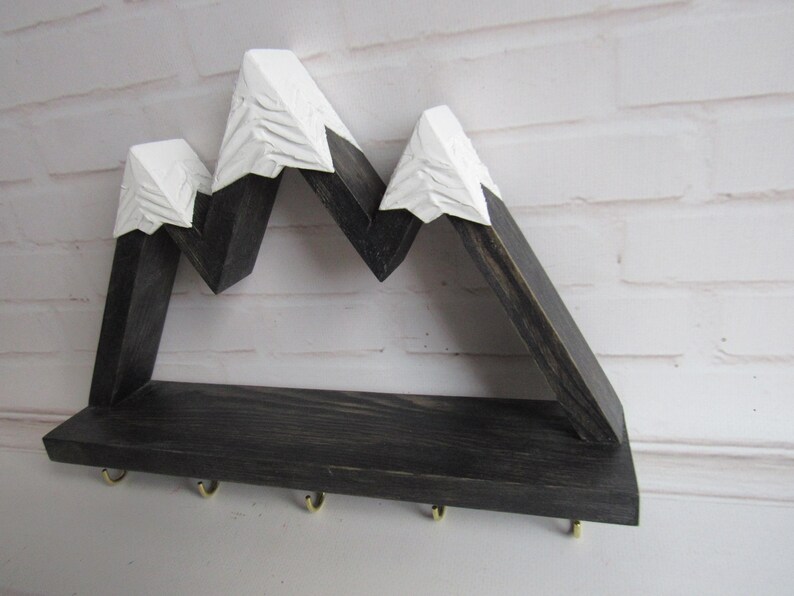 May include: A wall-mounted wooden shelf featuring a mountain range silhouette with dark charcoal stained wood bases and white painted peaks. The shelf includes five small gold-toned metal hooks attached to the bottom edge for hanging items.