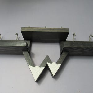 May include: A dark stained wooden wall shelf featuring three distinct levels and four silver metal hooks. The lower section includes two triangular supports painted white at the tips, creating a mountain-like silhouette with visible wood grain.