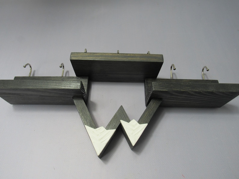 May include: A dark stained wooden wall shelf featuring three distinct levels and four silver metal hooks. The lower section includes two triangular supports painted white at the tips, creating a mountain-like silhouette with visible wood grain.