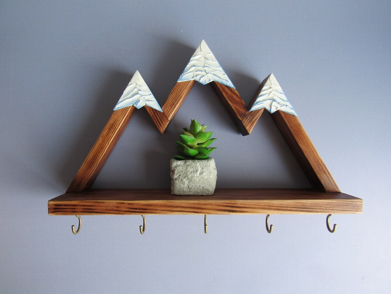 May include: Wooden mountain-shaped shelf with a small potted succulent plant in the center. The shelf has five hooks on the bottom and a white painted snow-capped mountain design.
