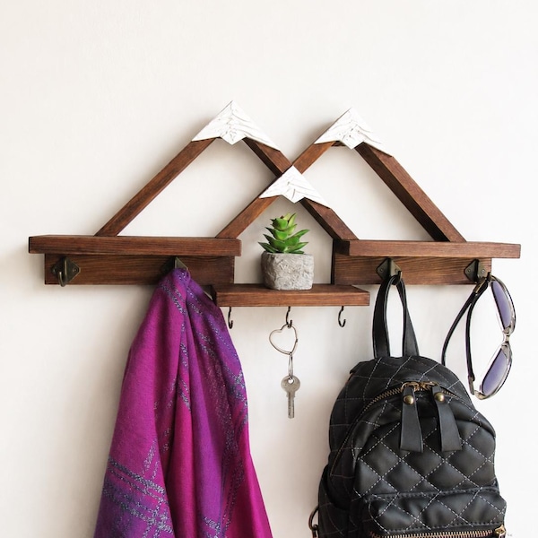 May include: A wall-mounted wooden shelf featuring a mountain-shaped frame with white peaks, three small horizontal platforms, and five metal hooks. A small artificial succulent in a gray pot sits on the center shelf, with a purple scarf, a black quilted backpack, a key, and sunglasses hanging from the hooks.