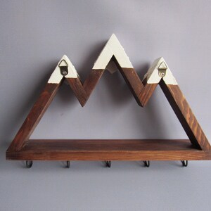 May include: A wall-mounted wooden shelf shaped like a mountain range with a dark brown stained finish and white painted peaks. The shelf features a flat base and five black metal hooks attached to the underside for hanging items, with two metal hangers.