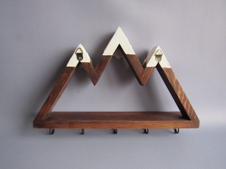 May include: A wall-mounted wooden shelf shaped like a mountain range with a dark brown stained finish and white painted peaks. The shelf features a flat base and five black metal hooks attached to the underside for hanging items, with two metal hangers.