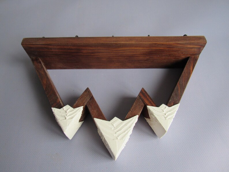 May include: A wall-mounted shelf featuring a dark brown stained wooden frame with a zigzag design. The three downward-pointing triangular peaks are finished with a thick, textured white paint, creating a mountain-like appearance on the lower edge.