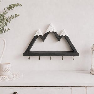 May include: A wall-mounted wooden shelf shaped like a mountain range with dark gray peaks and white snow-capped tops. The shelf features a flat horizontal base with five small metal hooks attached to the bottom edge for hanging various items.