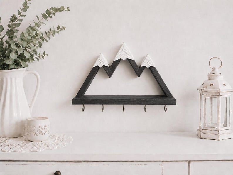 May include: A wall-mounted wooden shelf shaped like a mountain range with dark gray peaks and white snow-capped tops. The shelf features a flat horizontal base with five small metal hooks attached to the bottom edge for hanging various items.