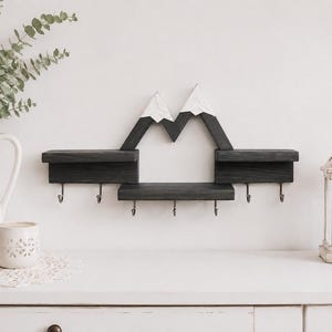 May include: A wall-mounted wooden shelf featuring a mountain-shaped design with white-painted peaks and dark gray stained wood. The unit includes three horizontal display surfaces and six metal hooks attached to the bottom for hanging items.