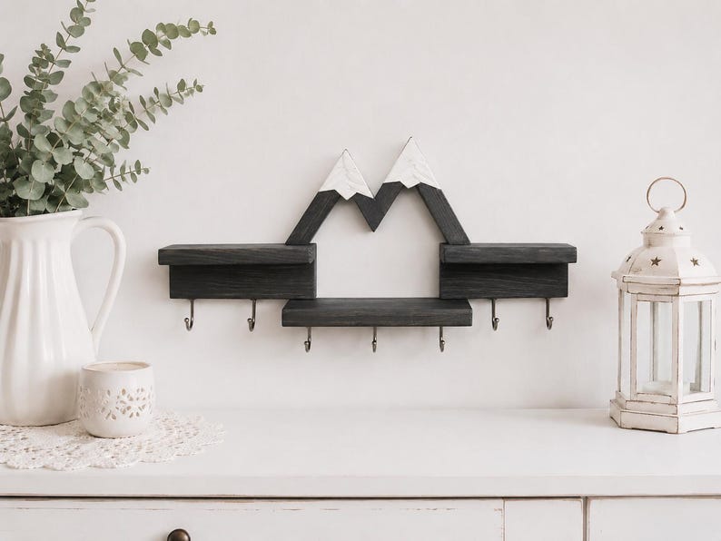 May include: A wall-mounted wooden shelf featuring a mountain-shaped design with white-painted peaks and dark gray stained wood. The unit includes three horizontal display surfaces and six metal hooks attached to the bottom for hanging items.