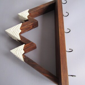 May include: A wall-mounted wooden rack shaped like a mountain range with dark brown stained wood and white painted peaks. The structure features four metal hooks attached to the straight vertical side for hanging items in a home or office space.