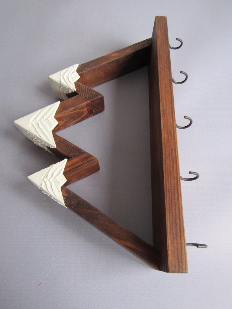 May include: A wall-mounted wooden rack shaped like a mountain range with dark brown stained wood and white painted peaks. The structure features four metal hooks attached to the straight vertical side for hanging items in a home or office space.