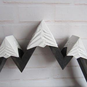 May include: A wooden mountain-shaped wall decor piece featuring three peaks. The lower sections are finished in a dark charcoal stain, while the upper portions are painted with a thick, textured white finish to resemble snow-capped summits.