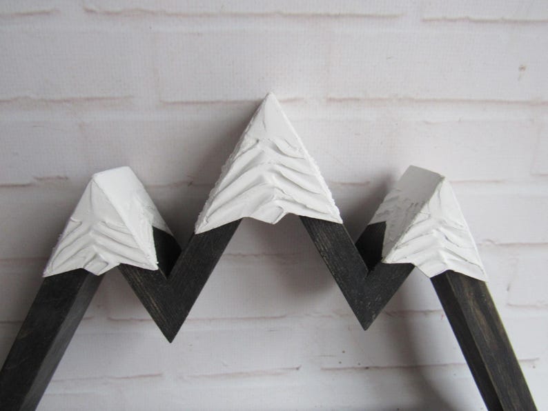 May include: A wooden mountain-shaped wall decor piece featuring three peaks. The lower sections are finished in a dark charcoal stain, while the upper portions are painted with a thick, textured white finish to resemble snow-capped summits.