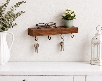 May include: A rectangular dark brown wooden wall shelf featuring four metal hooks along the bottom edge. Two sets of silver-toned keys hang from the hooks. The top surface of the shelf holds a pair of dark-rimmed eyeglasses.