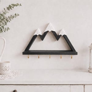 May include: A wall-mounted wooden shelf shaped like a mountain range with black peaks and white snow-capped tops. The shelf features a flat black base with five small gold-colored metal hooks attached to the bottom edge for hanging items.