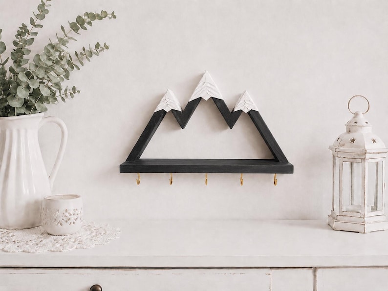 May include: A wall-mounted wooden shelf shaped like a mountain range with black peaks and white snow-capped tops. The shelf features a flat black base with five small gold-colored metal hooks attached to the bottom edge for hanging items.