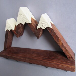 May include: A wall-mounted wooden shelf shaped like a mountain range with three peaks. The base and lower mountain sections are made of dark brown stained wood, while the pointed tops are painted in an off-white color with carved textures.