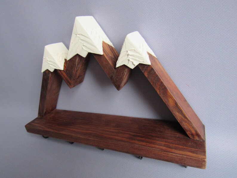May include: A wall-mounted wooden shelf shaped like a mountain range with three peaks. The base and lower mountain sections are made of dark brown stained wood, while the pointed tops are painted in an off-white color with carved textures.