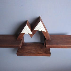 May include: A wall-mounted wooden shelf unit featuring a dark brown finish and a mountain-shaped design. The structure includes three flat horizontal surfaces for storage and two white triangular peaks at the top with carved line details.