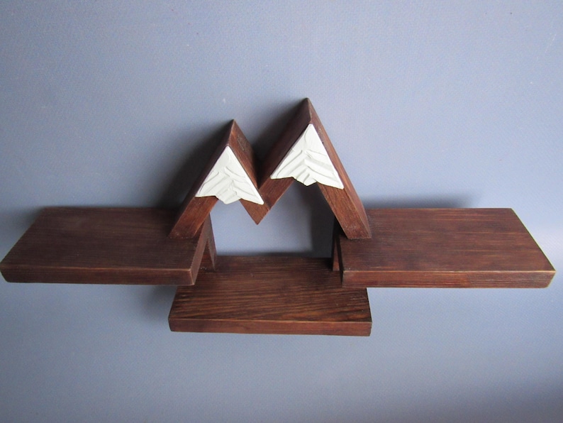 May include: A wall-mounted wooden shelf unit featuring a dark brown finish and a mountain-shaped design. The structure includes three flat horizontal surfaces for storage and two white triangular peaks at the top with carved line details.