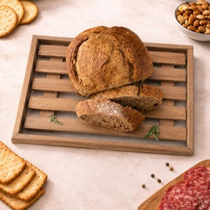 May include: A rectangular wooden bread board with a slatted design, featuring a large round loaf of crusty brown bread with two slices cut and placed on top, accompanied by small sprigs of green rosemary resting on the wooden slats.
