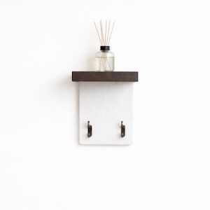 May include: A small, wall-mounted shelf with a dark brown top and a white base. Two bronze-coloured hooks are attached to the base. A glass bottle with reeds sits on the shelf, suggesting a home fragrance product.