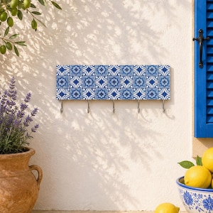 Pode incluir: Um cabide de parede retangular com um padrão de azulejos geométricos repetitivos em azul e branco. A placa horizontal inclui cinco ganchos de metal prateado espaçados uniformemente na borda inferior para pendurar objetos.
