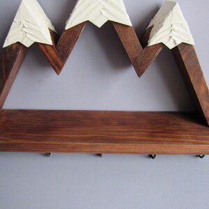 May include: A wall-mounted wooden shelf shaped like a mountain range with three peaks. The structure is made of dark brown stained wood with textured, off-white painted tips on each peak. Five small black metal hooks are attached to the bottom edge.