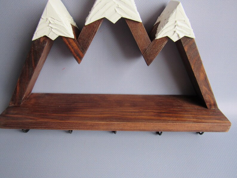 May include: A wall-mounted wooden shelf shaped like a mountain range with three peaks. The structure is made of dark brown stained wood with textured, off-white painted tips on each peak. Five small black metal hooks are attached to the bottom edge.