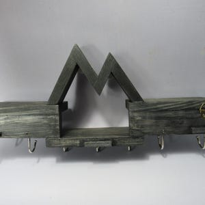 May include: A dark gray stained wooden wall shelf featuring a mountain-shaped silhouette. The unit includes two flat storage surfaces, a central lower shelf, and seven silver-toned metal hooks attached to the bottom for hanging items.